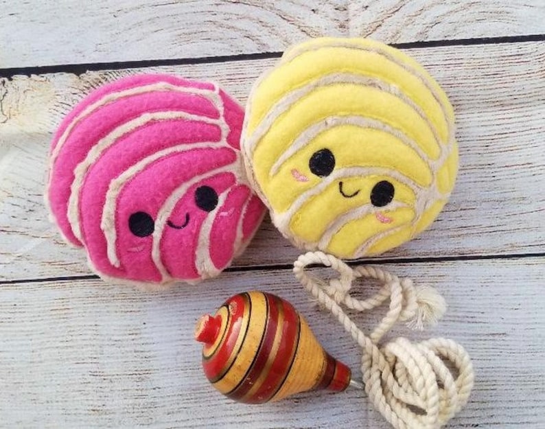 Pu&ograve; includere: Due giocattoli di peluche a forma di pane dolce messicano, uno rosa e uno giallo, con facce sorridenti. Una trottola di legno con strisce rosse e gialle &egrave; in primo piano.