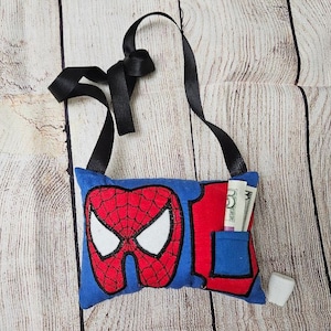 May include: A blue and red tooth pillow with a Spiderman design. The pillow has a pocket on the side for holding money or a tooth. The pillow is attached to a black ribbon.