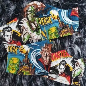 May include: Two fabric face masks with a horror theme. The masks feature a blue background with images of monsters, including Frankenstein, Dracula, and a werewolf. The text "Horror" and "Monster" are printed on the masks.
