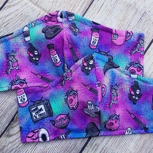 May include: Two purple and blue face masks with a variety of colorful and black graphic designs. The designs include a TV with the text "True Crime & Chills", a skull, a syringe, a bottle of pills, a ghost, and a coffee mug.