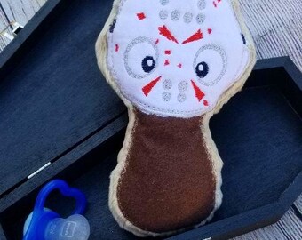 May include: A plush toy shaped like a hockey mask with red accents and a brown body, placed inside a black coffin-shaped box. A blue pacifier is also in the box. The toy has a white face with red details.