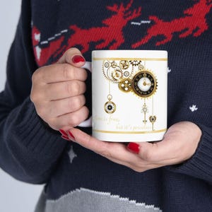 May include: White ceramic mug with a gold and black clockwork design, including gears, clocks, and keys. The mug has the text "Time is free, but it's priceless" in gold script. The mug is held by a person wearing a blue sweater.