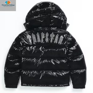 May include: Black puffer jacket with a hood. The jacket has a shiny, glossy finish and features the word "TRAPSTAR" in a stylized font across the upper back. The jacket is designed with horizontal quilted panels.