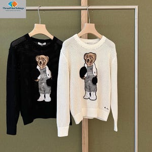May include: Two sweaters hanging on a rack. One is black, the other is off-white. Both have a bear graphic wearing a gray outfit and boxing gloves. The sweaters are made of a knit material.