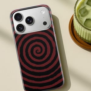 May include: A clear phone case with a red and black spiral design. The case has a camera cutout with three lenses and a flash. The phone case is on a light beige surface.