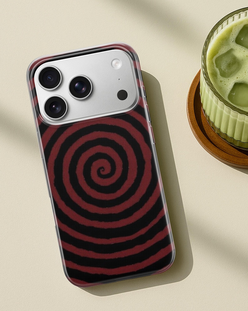 May include: A clear phone case with a red and black spiral design. The case has a camera cutout with three lenses and a flash. The phone case is on a light beige surface.