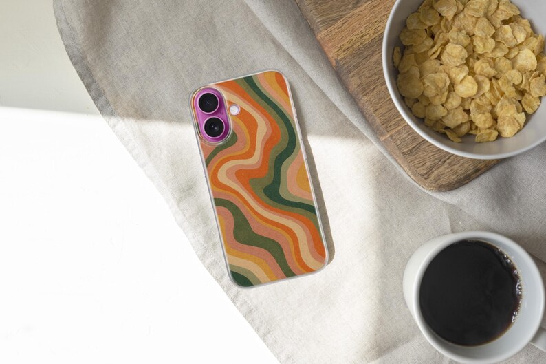 Pode incluir: Uma capa de smartphone com um padr&atilde;o ondulado colorido em tons de laranja, verde e bege. A capa est&aacute; sobre uma superf&iacute;cie clara, perto de uma tigela de cereais e uma x&iacute;cara de caf&eacute;.