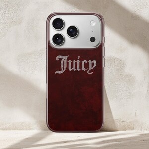 May include: A burgundy phone case with the word "Juicy" in a rhinestone-style font. The case has a dark red, textured back and a clear border. The phone's camera lenses are visible at the top.