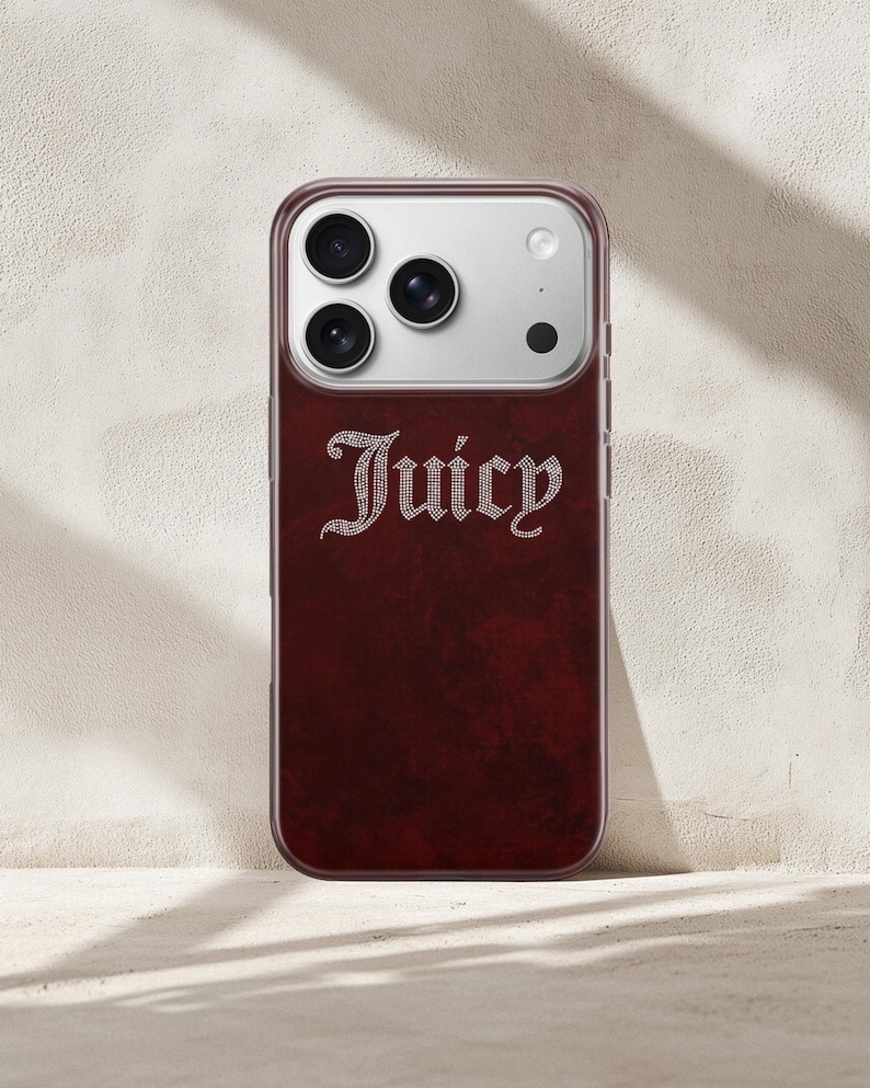 May include: A burgundy phone case with the word "Juicy" in a rhinestone-style font. The case has a dark red, textured back and a clear border. The phone's camera lenses are visible at the top.