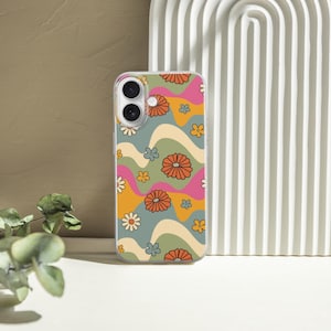 Pode incluir: Uma capa de telem&oacute;vel com um design floral retro. A capa apresenta um padr&atilde;o de flores laranja, rosa e brancas sobre um fundo ondulado de verde, amarelo e azul. A capa &eacute; mostrada numa superf&iacute;cie branca com um arco decorativo e vegeta&ccedil;&atilde;o.