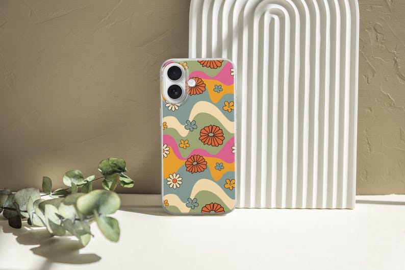 Pode incluir: Uma capa de telem&oacute;vel com um design floral retro. A capa apresenta um padr&atilde;o de flores laranja, rosa e brancas sobre um fundo ondulado de verde, amarelo e azul. A capa &eacute; mostrada numa superf&iacute;cie branca com um arco decorativo e vegeta&ccedil;&atilde;o.