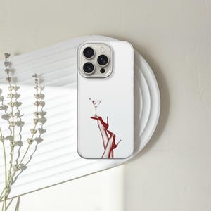 Puede incluir: Funda de tel&eacute;fono blanca con una ilustraci&oacute;n roja de una copa de martini equilibrada sobre un par de tacones rojos. La funda est&aacute; sobre un plato blanco, con ramitas de lavanda seca a la izquierda.