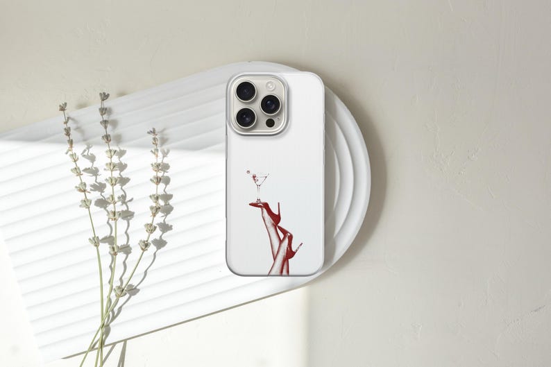 Puede incluir: Funda de tel&eacute;fono blanca con una ilustraci&oacute;n roja de una copa de martini equilibrada sobre un par de tacones rojos. La funda est&aacute; sobre un plato blanco, con ramitas de lavanda seca a la izquierda.
