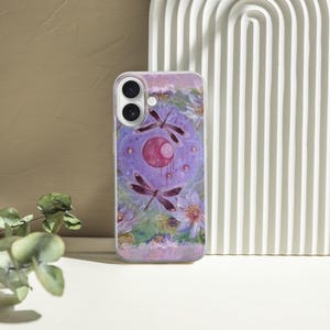May include: A lavender phone case featuring a watercolor design with dragonflies, a crescent moon, and floral elements. The case has a glossy finish and a camera cutout. The background includes a white arched structure and a textured wall.