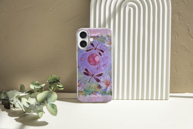 May include: A lavender phone case featuring a watercolor design with dragonflies, a crescent moon, and floral elements. The case has a glossy finish and a camera cutout. The background includes a white arched structure and a textured wall.