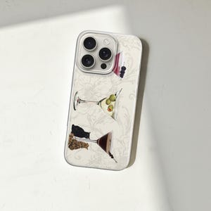 Puede incluir: Funda de tel&eacute;fono con un estampado floral blanco e ilustraciones de copas de martini. Las copas de martini contienen aceitunas, bayas y otras guarniciones. La funda tiene un protector de lente de c&aacute;mara transparente.