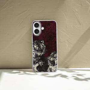 May include: A clear phone case with a burgundy background featuring a black and white illustration of three roaring tigers. The case has a white border and is designed for a smartphone with dual cameras.