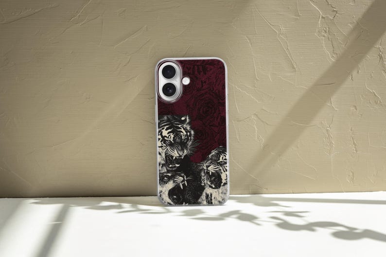 May include: A clear phone case with a burgundy background featuring a black and white illustration of three roaring tigers. The case has a white border and is designed for a smartphone with dual cameras.