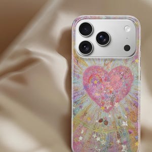 May include: A clear phone case featuring a colorful design with a large pink heart at the center. The heart is surrounded by a burst of light and stars, with small strawberries and glitter accents. The case has a clear border and covers the phone's back.