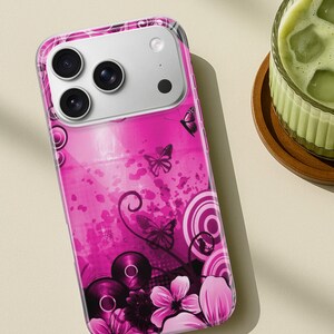 May include: A pink phone case with a butterfly and floral design. The case features a clear border and a vibrant pink background with abstract shapes, butterflies, and flowers. The phone's camera lenses are visible at the top.