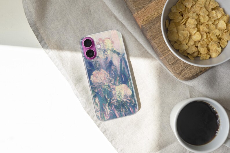 May include: A smartphone case with a floral design in shades of pink, purple, and blue. The case is on a light-colored surface, next to a bowl of cereal and a cup of coffee. The phone case has a pink camera module.