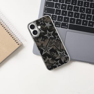 May include: A smartphone case with a black and gray star pattern rests on a laptop. A spiral-bound notebook with a brown cover is to the left. The laptop is open on a white surface.