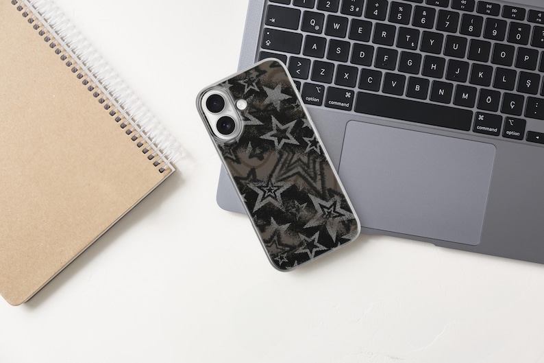 May include: A smartphone case with a black and gray star pattern rests on a laptop. A spiral-bound notebook with a brown cover is to the left. The laptop is open on a white surface.