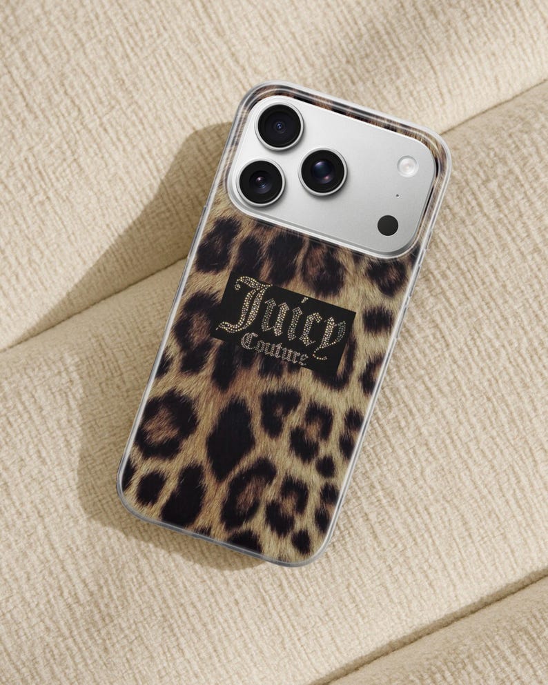 May include: A clear phone case with a leopard print design. The case features the text "Juicy Couture" in a black rectangle with a rhinestone effect. The phone case is on a beige surface.