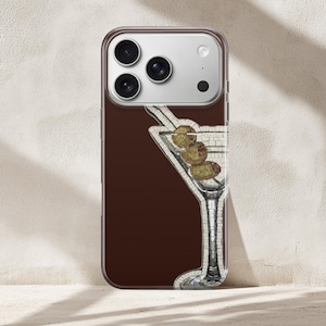Puede incluir: Una funda de tel&eacute;fono con un fondo marr&oacute;n, que muestra una copa de martini de estilo mosaico con aceitunas. La funda tiene un borde transparente y un m&oacute;dulo de c&aacute;mara.