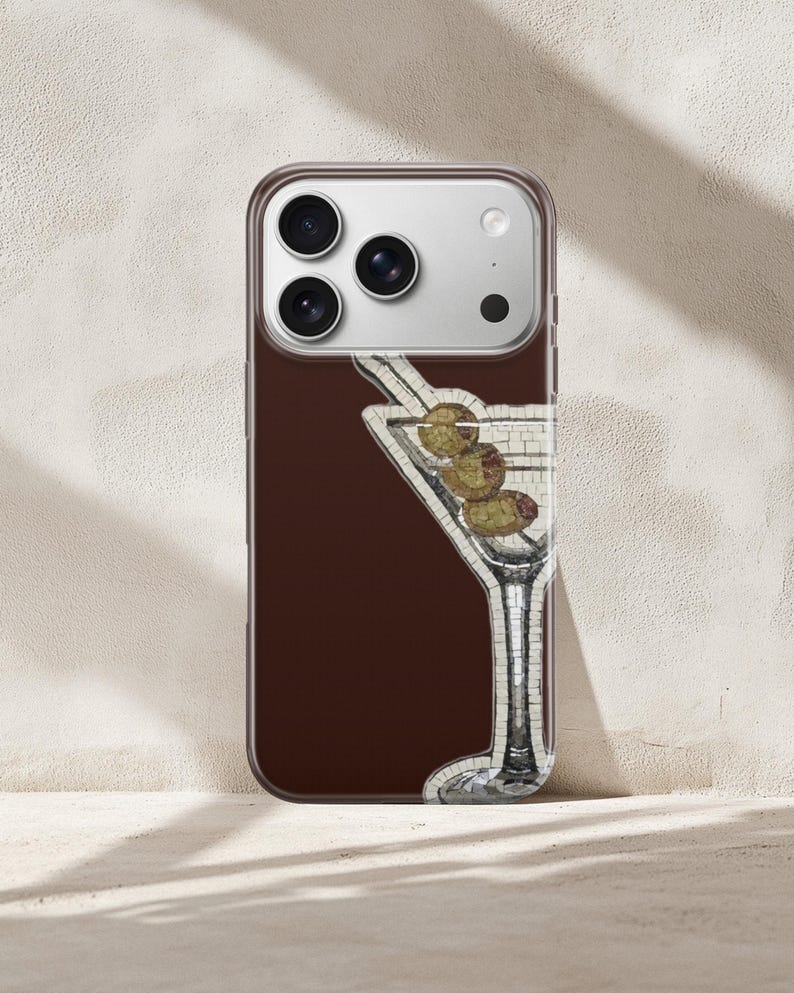 Puede incluir: Una funda de tel&eacute;fono con un fondo marr&oacute;n, que muestra una copa de martini de estilo mosaico con aceitunas. La funda tiene un borde transparente y un m&oacute;dulo de c&aacute;mara.
