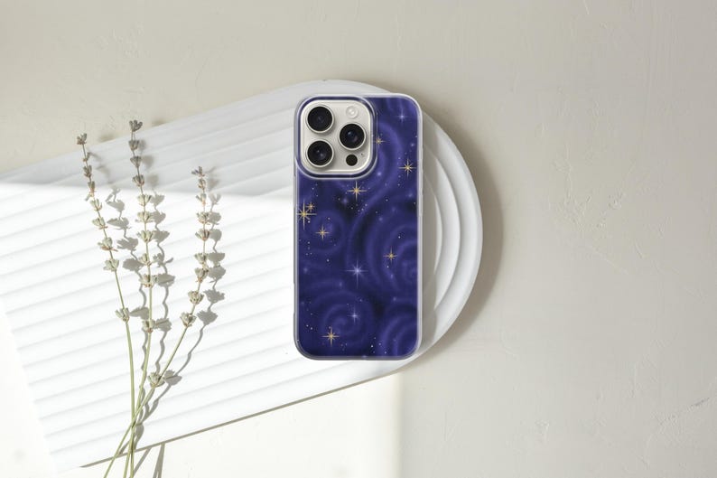 May include: A smartphone case with a dark blue, swirling galaxy design, featuring gold stars. The case is on a white, circular plate, with dried lavender sprigs to the left. The case has a clear border.