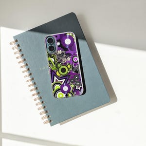 May include: A smartphone case with a colorful retro design featuring purple, green, and white circles, stars, and floral elements. The case rests on a blue spiral-bound notebook with gold-colored coils. The notebook is open to a page with the word "TWO" visible.
