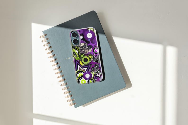 May include: A smartphone case with a colorful retro design featuring purple, green, and white circles, stars, and floral elements. The case rests on a blue spiral-bound notebook with gold-colored coils. The notebook is open to a page with the word "TWO" visible.