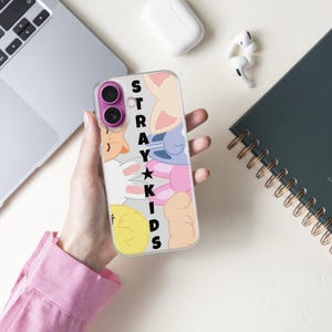 May include: A clear phone case featuring a colorful cartoon design with the text "STRAY KIDS" in black. The case is held in a hand, with a laptop, notebook, and wireless earbuds in the background.