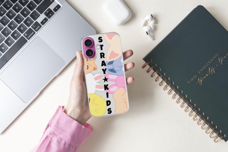 May include: A clear phone case featuring a colorful cartoon design with the text "STRAY KIDS" in black. The case is held in a hand, with a laptop, notebook, and wireless earbuds in the background.