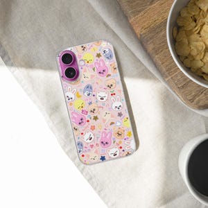 May include: A clear phone case with a pink and white pattern featuring cartoon animal faces and stars. The case has a dual-lens camera cutout with a pink ring. The phone case is on a light-colored surface.