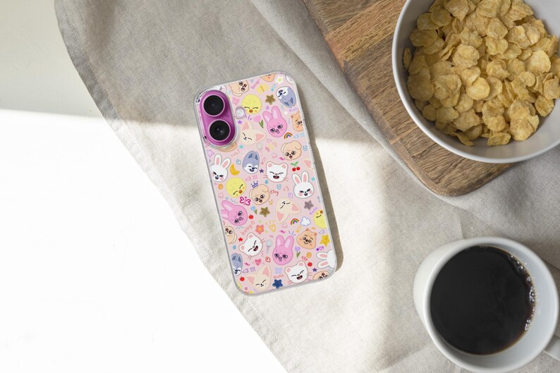 May include: A clear phone case with a pink and white pattern featuring cartoon animal faces and stars. The case has a dual-lens camera cutout with a pink ring. The phone case is on a light-colored surface.