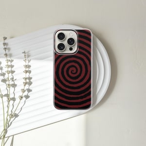 May include: A smartphone case with a red and black spiral design. The case is on a white plate with a textured surface. Dried flowers are to the left of the plate. The phone case has a clear border.