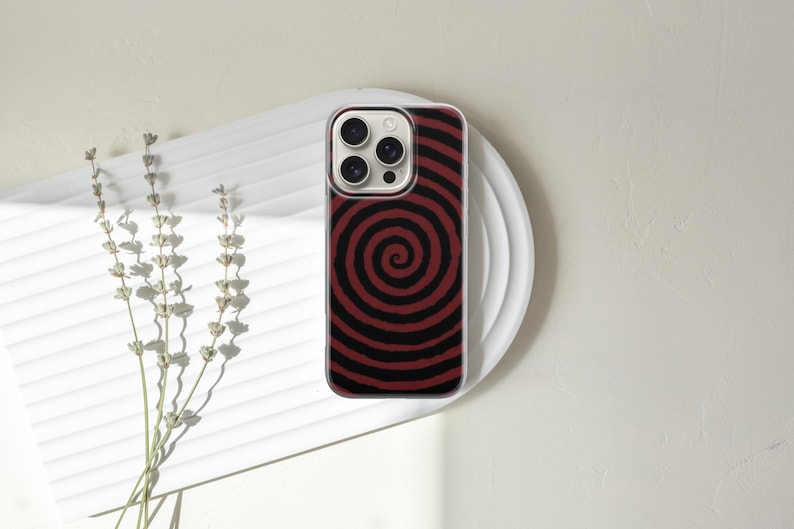 May include: A smartphone case with a red and black spiral design. The case is on a white plate with a textured surface. Dried flowers are to the left of the plate. The phone case has a clear border.