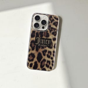 May include: A phone case with a leopard print design. The case features a brown and black spotted pattern, with the words "Juicy Couture" in a black rectangular box. The case has a clear border and a camera cutout.