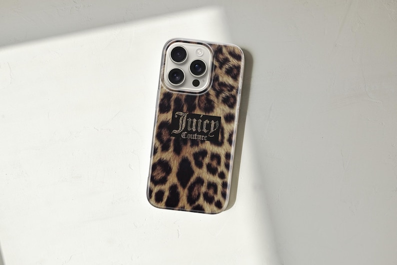 May include: A phone case with a leopard print design. The case features a brown and black spotted pattern, with the words "Juicy Couture" in a black rectangular box. The case has a clear border and a camera cutout.