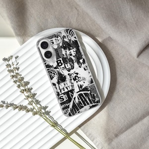 May include: A black and white patterned phone case with a collage design, resting on a white plate. The case features a mix of text and abstract shapes. A sprig of dried flowers is placed near the plate, with a neutral fabric in the background.