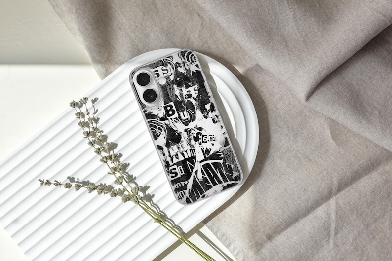 May include: A black and white patterned phone case with a collage design, resting on a white plate. The case features a mix of text and abstract shapes. A sprig of dried flowers is placed near the plate, with a neutral fabric in the background.