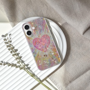 May include: A clear phone case featuring a pink heart design with a sparkling, colorful background. The case is on a white, ridged surface, with a sprig of dried lavender to the side. The phone case is a protective accessory.