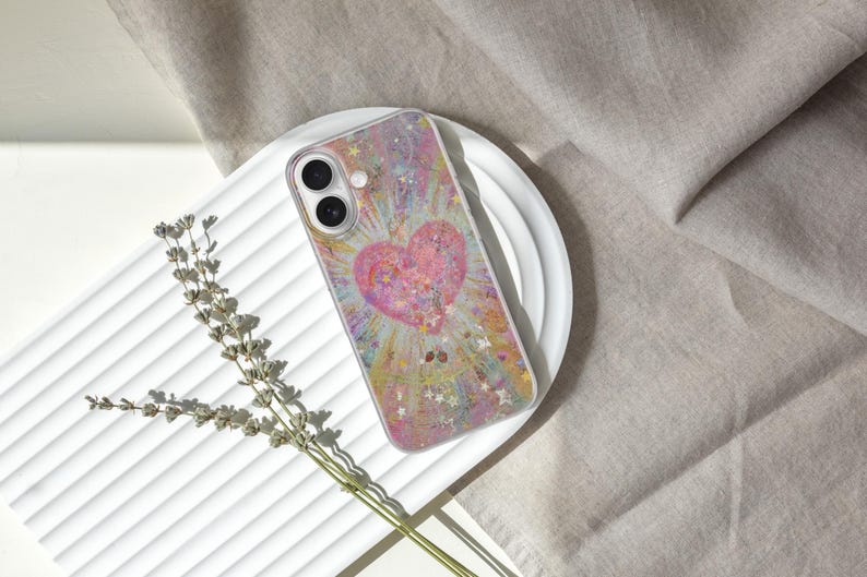 May include: A clear phone case featuring a pink heart design with a sparkling, colorful background. The case is on a white, ridged surface, with a sprig of dried lavender to the side. The phone case is a protective accessory.