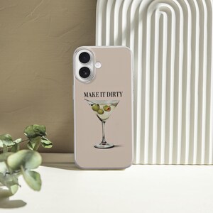 Puede incluir: Funda de tel&eacute;fono beige con un dise&ntilde;o de copa de Martini, aceitunas y el texto "MAKE IT DIRTY". El fondo presenta una estructura arqueada blanca y follaje.