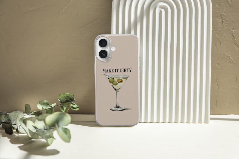 Puede incluir: Funda de tel&eacute;fono beige con un dise&ntilde;o de copa de Martini, aceitunas y el texto "MAKE IT DIRTY". El fondo presenta una estructura arqueada blanca y follaje.