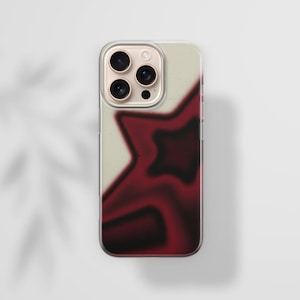 May include: A clear phone case with a cream-colored background and a blurred, dark red star design. The phone case has a gold-colored camera module. The case is on a white surface.
