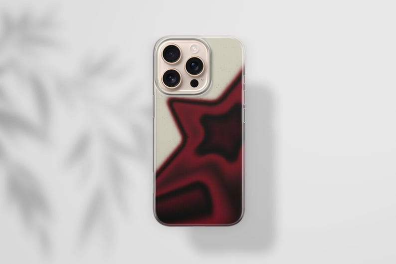 May include: A clear phone case with a cream-colored background and a blurred, dark red star design. The phone case has a gold-colored camera module. The case is on a white surface.