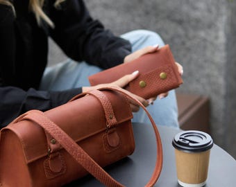 May include: A brown leather handbag with a long strap and a matching wallet with gold-colored button closures. The handbag is cylindrical and sits on a black table next to a disposable coffee cup.